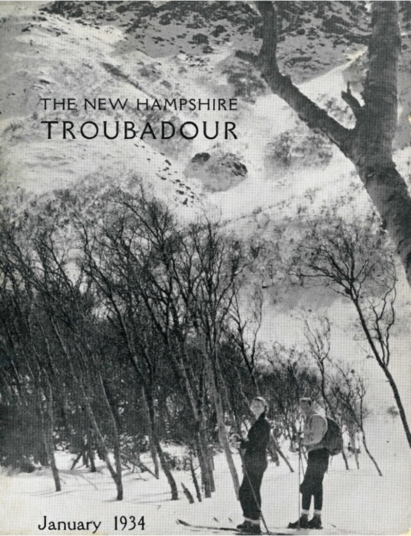 Two skiers with poles and rucksacks stand among bare winter trees on a snowy slope, with a snow-covered mountainside visible above them.